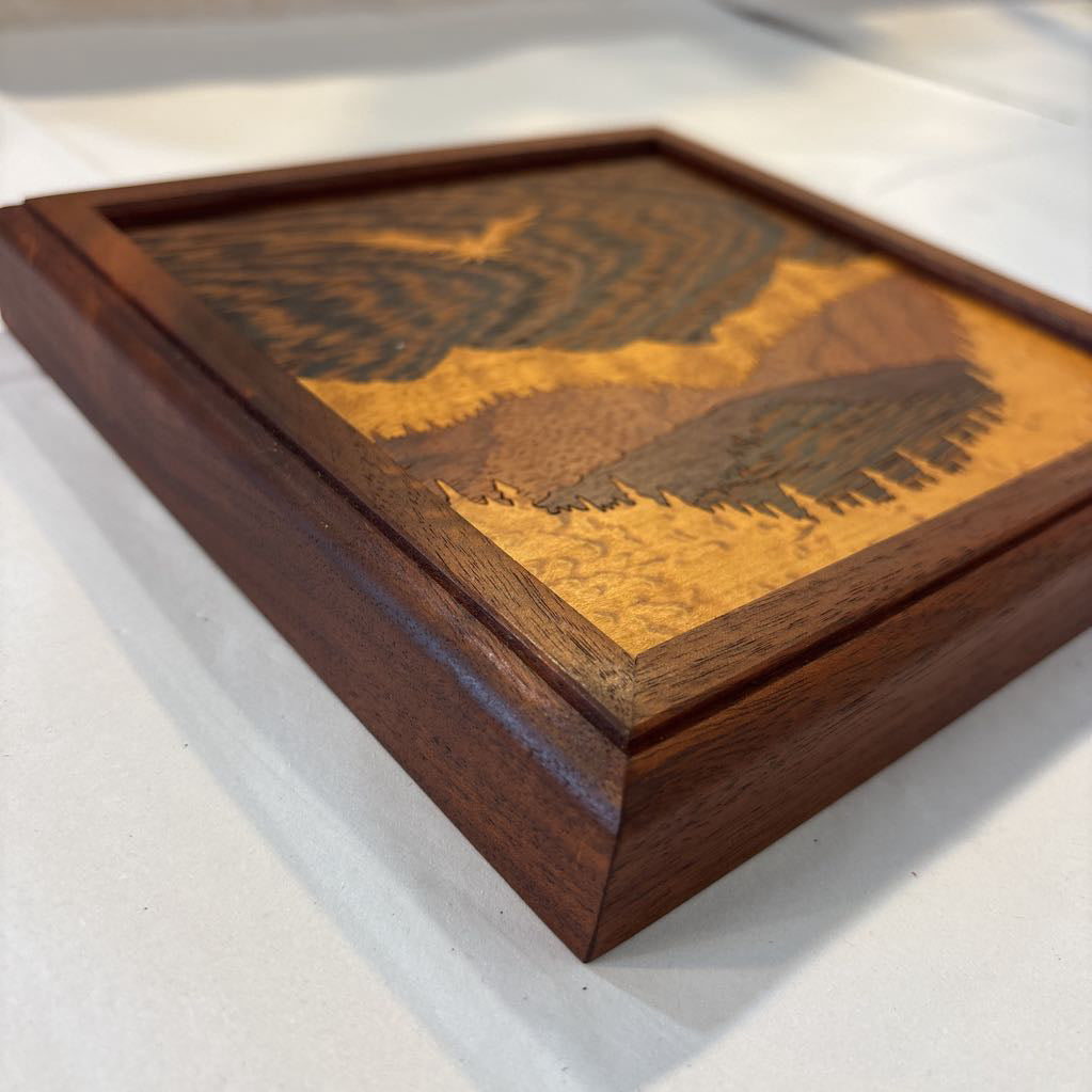A square framed wooden inlaid artwork of a bird flying over mountains and forests with different colors and textures of wood. 