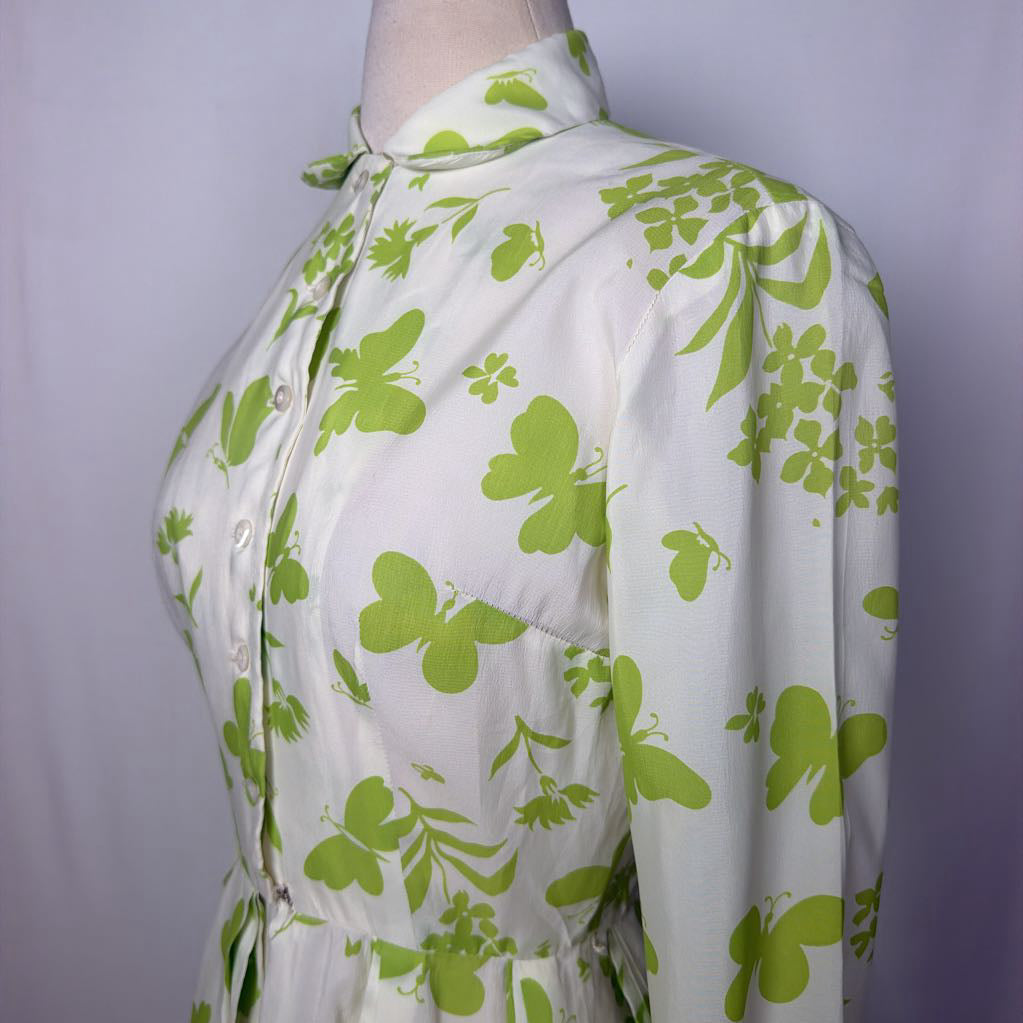 A long-sleeve collared dress displayed on a mannequin torso against a light gray fabric backdrop. The dress has an off-white base with a bright green floral and leaf print, a button front, and a self-tie waist belt.