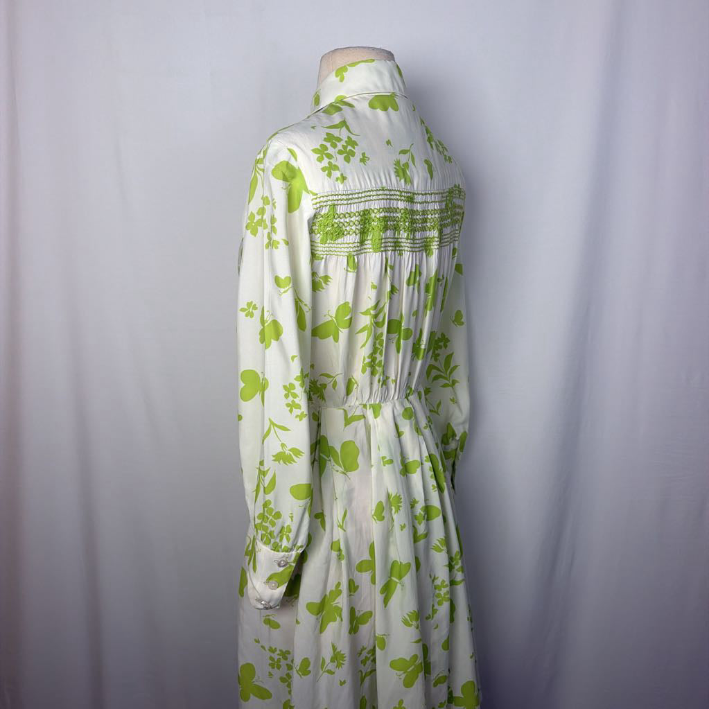 A long-sleeve white dress with a green floral and leaf pattern, featuring smocked detailing across the chest, displayed on a dress form against a light gray fabric backdrop.