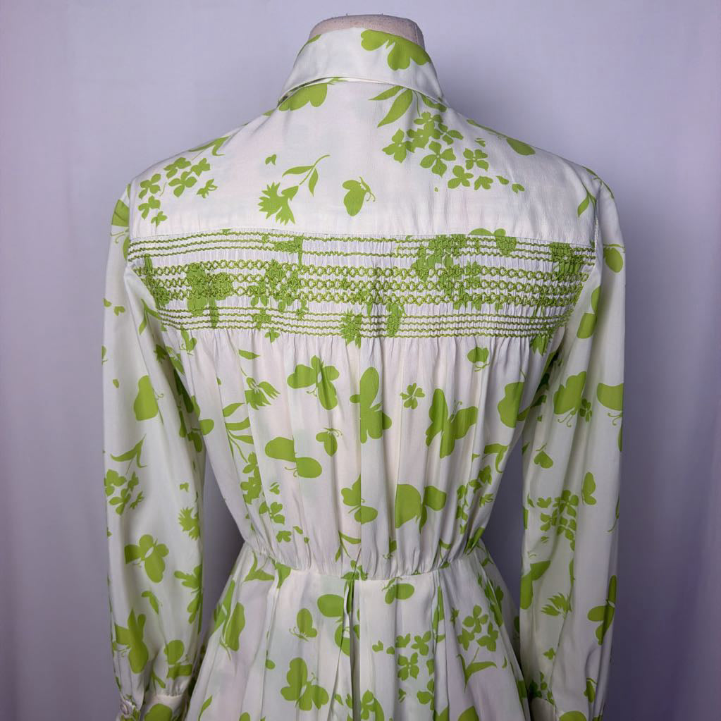 A long-sleeve collared dress shown from the back on a mannequin, featuring a white base with a green floral and leaf pattern and a smocked horizontal band across the upper back.