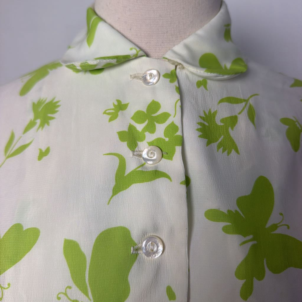 A close-up of a light green and white floral print blouse displayed on a mannequin torso, featuring a collar and three clear plastic snap buttons.
