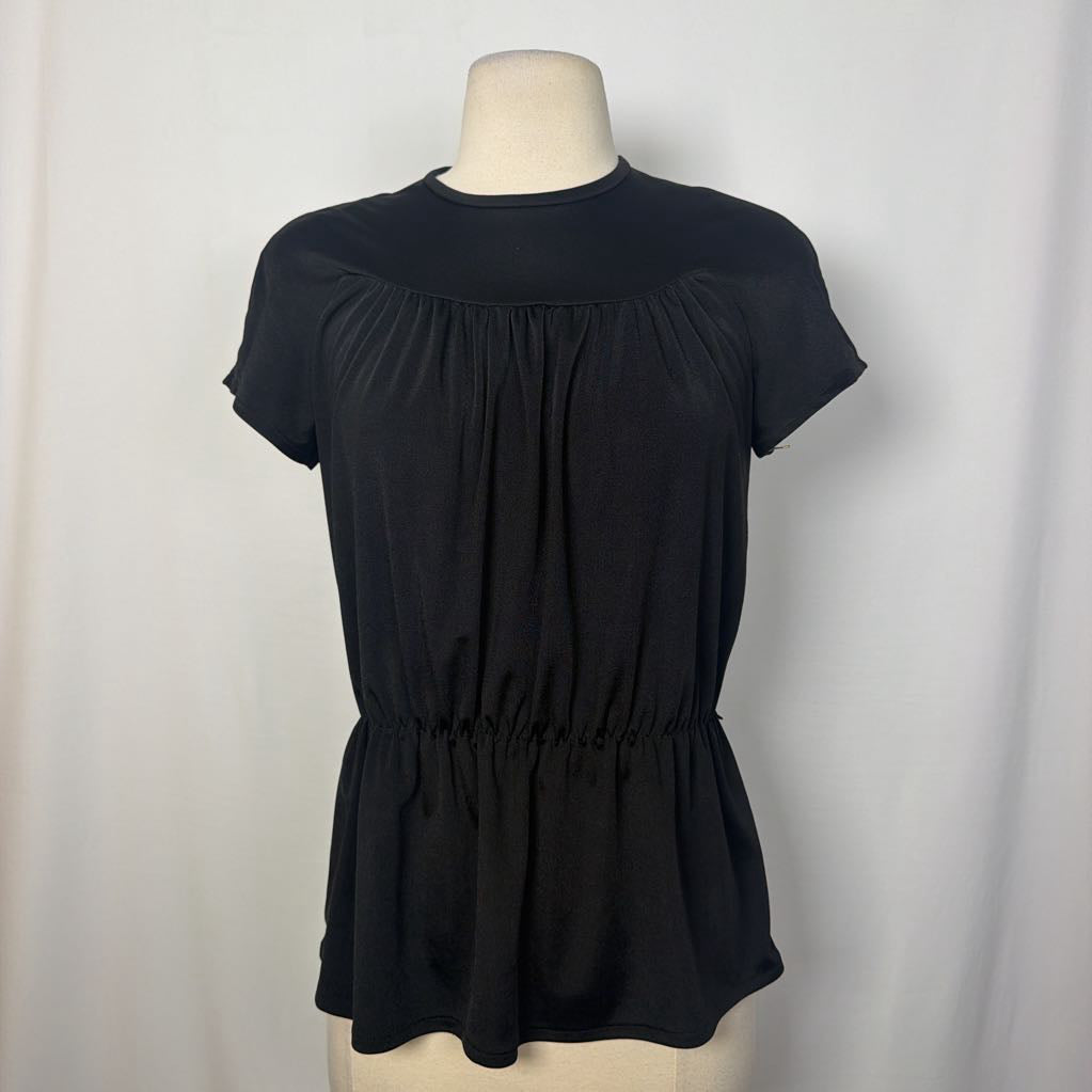 A black short-sleeve top with a gathered peplum waist, displayed on a cream mannequin torso against a white fabric backdrop.