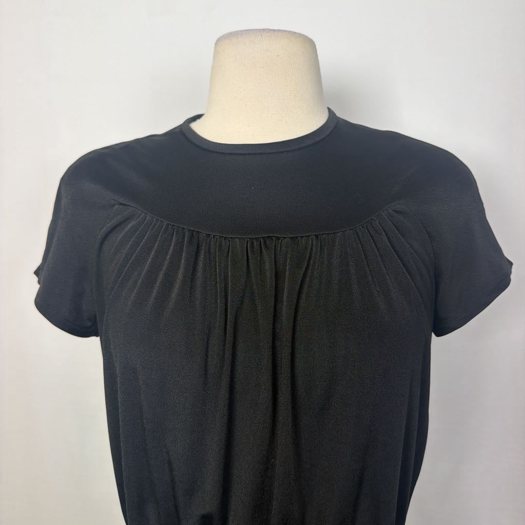 A black short-sleeve top with a gathered, pleated front bodice and a wide crew neckline, displayed on a beige mannequin torso against a light gray background.