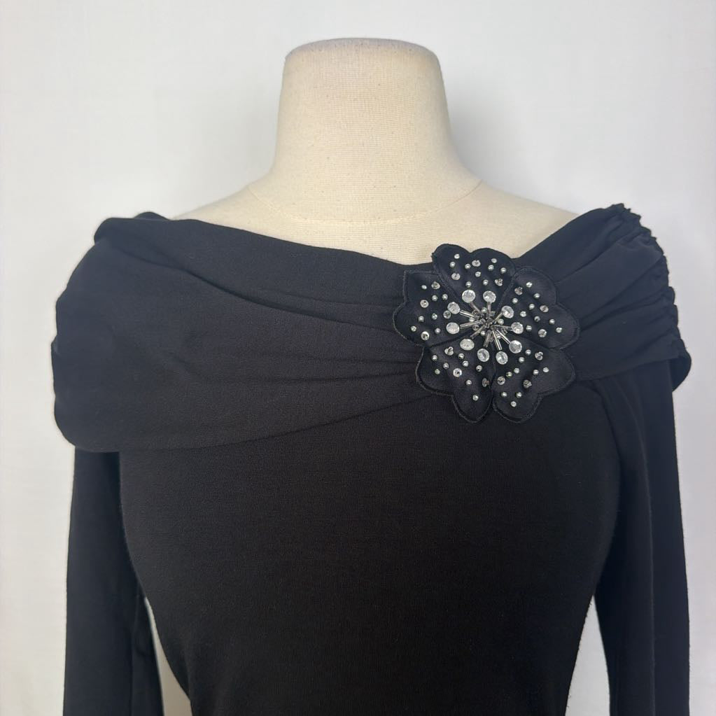 A black top with 3/4 sleeves and a draped collar with a large flower adornment on a mannequin in front of a white wall. 