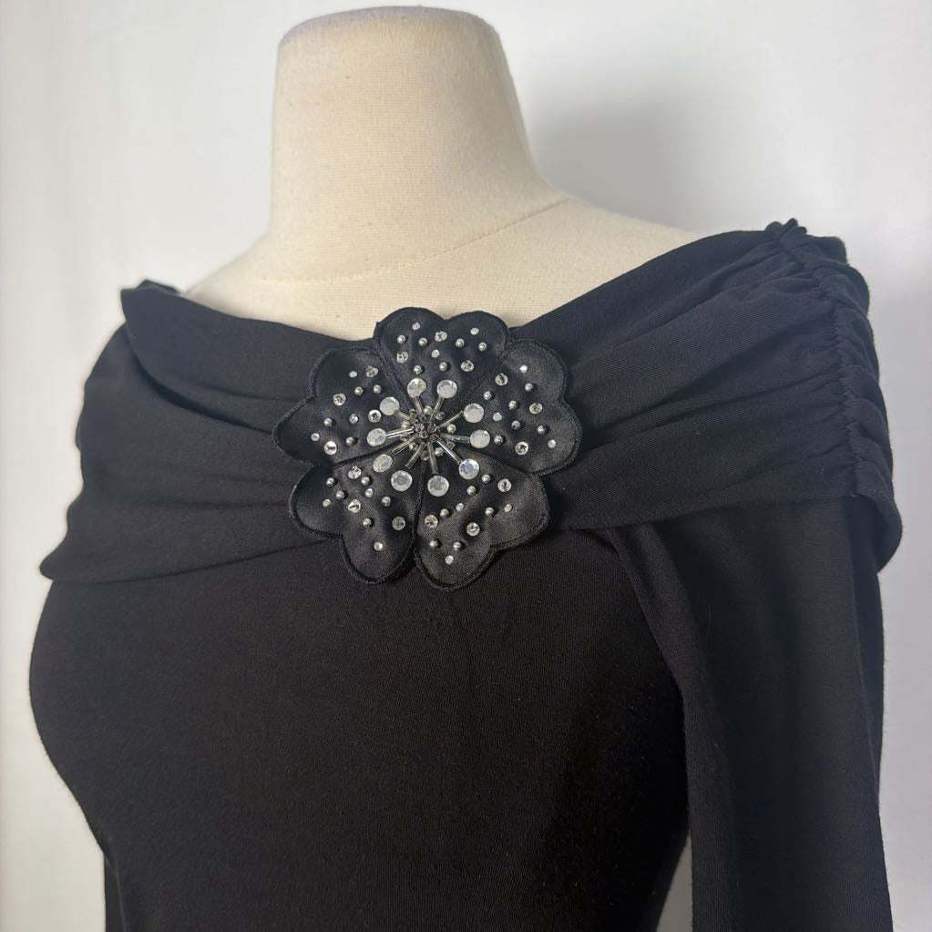 A black top with 3/4 sleeves and a draped collar with a large flower adornment on a mannequin in front of a white wall. 