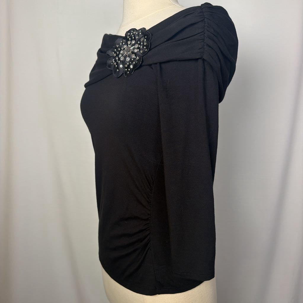 A black top with 3/4 sleeves and a draped collar with a large flower adornment on a mannequin in front of a white wall. 