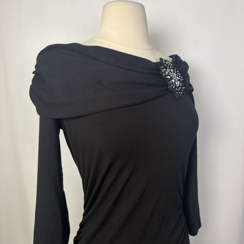 A black top with 3/4 sleeves and a draped collar with a large flower adornment on a mannequin in front of a white wall. 