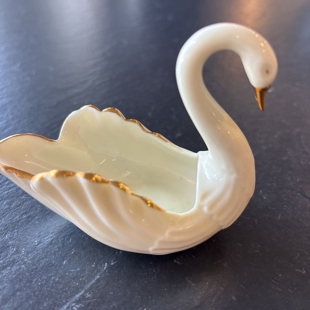 A small ceramic white swan figurine with gold trim on the wings and beak on a dark surface. 
