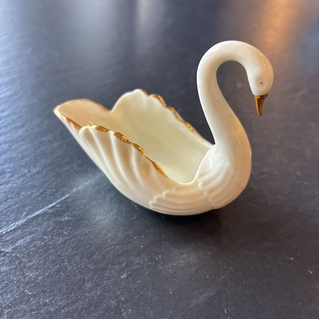 A small ceramic white swan figurine with gold trim on the wings and beak on a dark surface. 