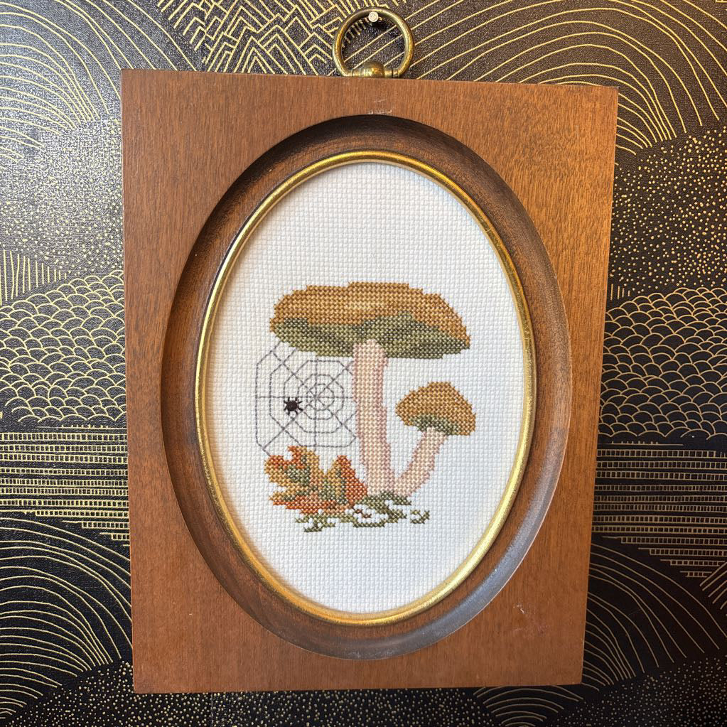 A mushroom and spider cross stitch artwork in a wooden frame hanging on a black and gold wall. 