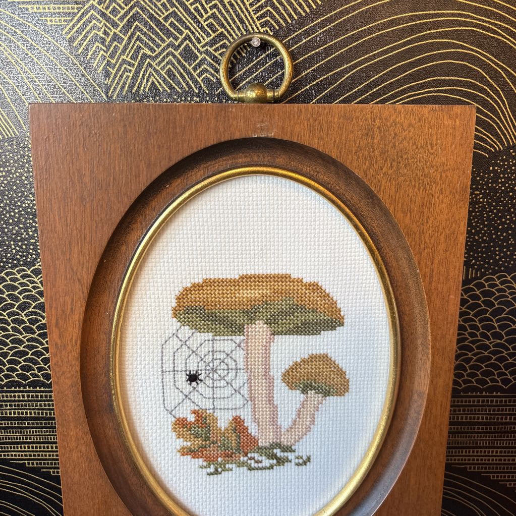 A mushroom and spider cross stitch artwork in a wooden frame hanging on a black and gold wall. 