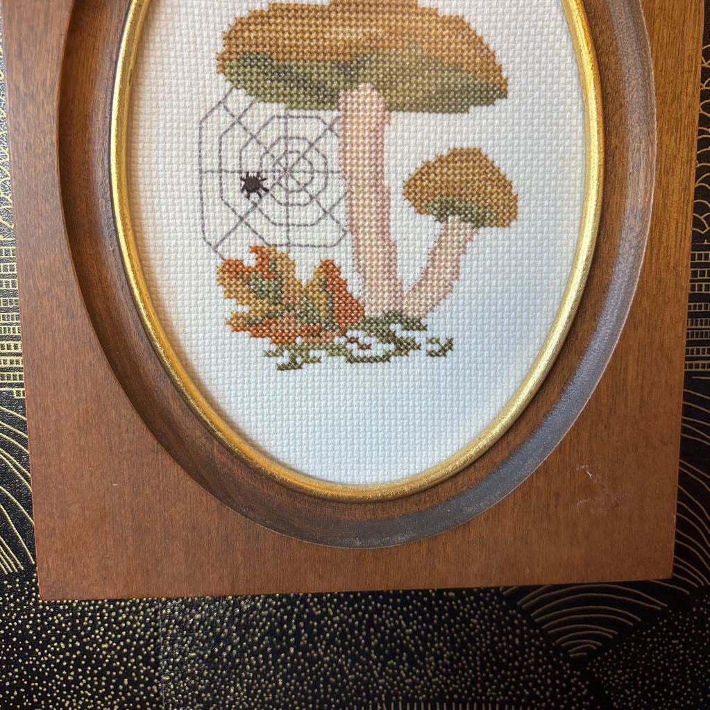A mushroom and spider cross stitch artwork in a wooden frame hanging on a black and gold wall. 