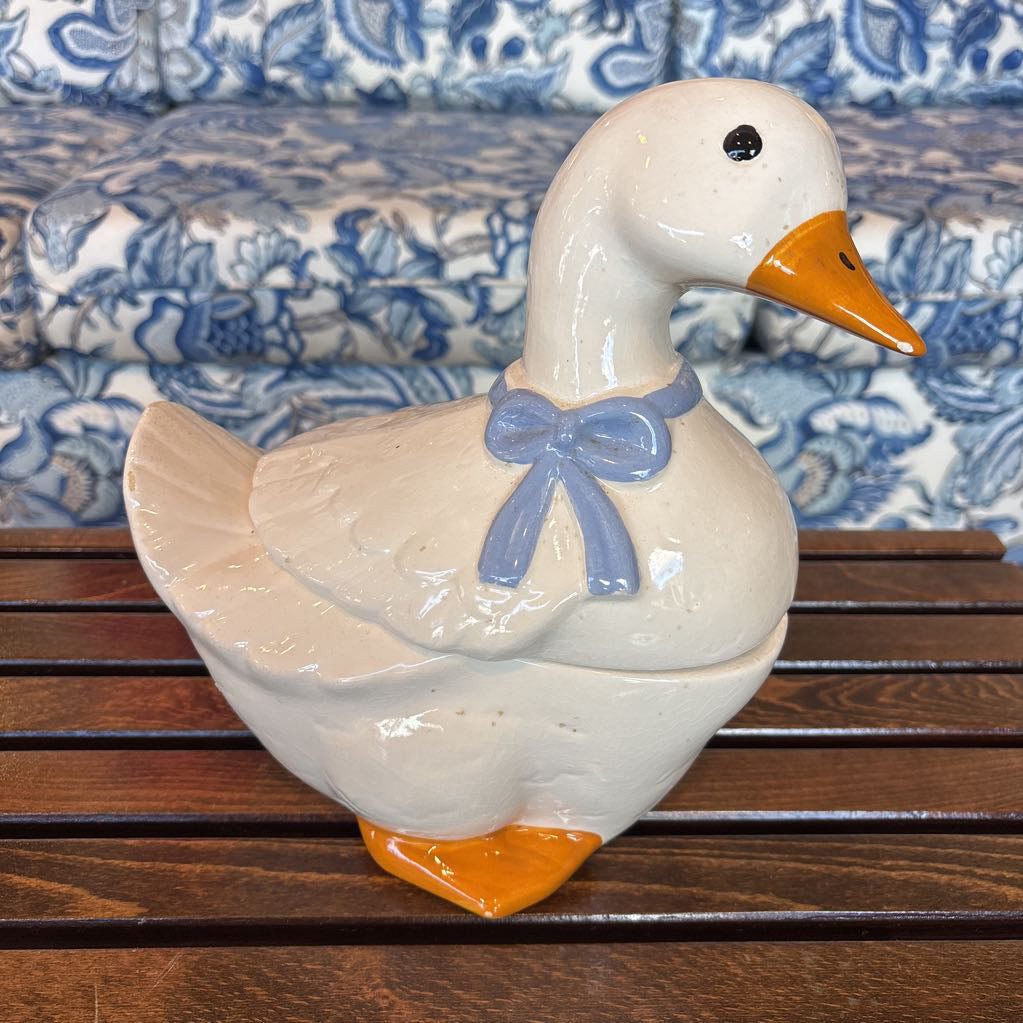 A ceramic white duck figure with a blue bow around its neck sitting on a wooden table in front of a blue and white sofa. 