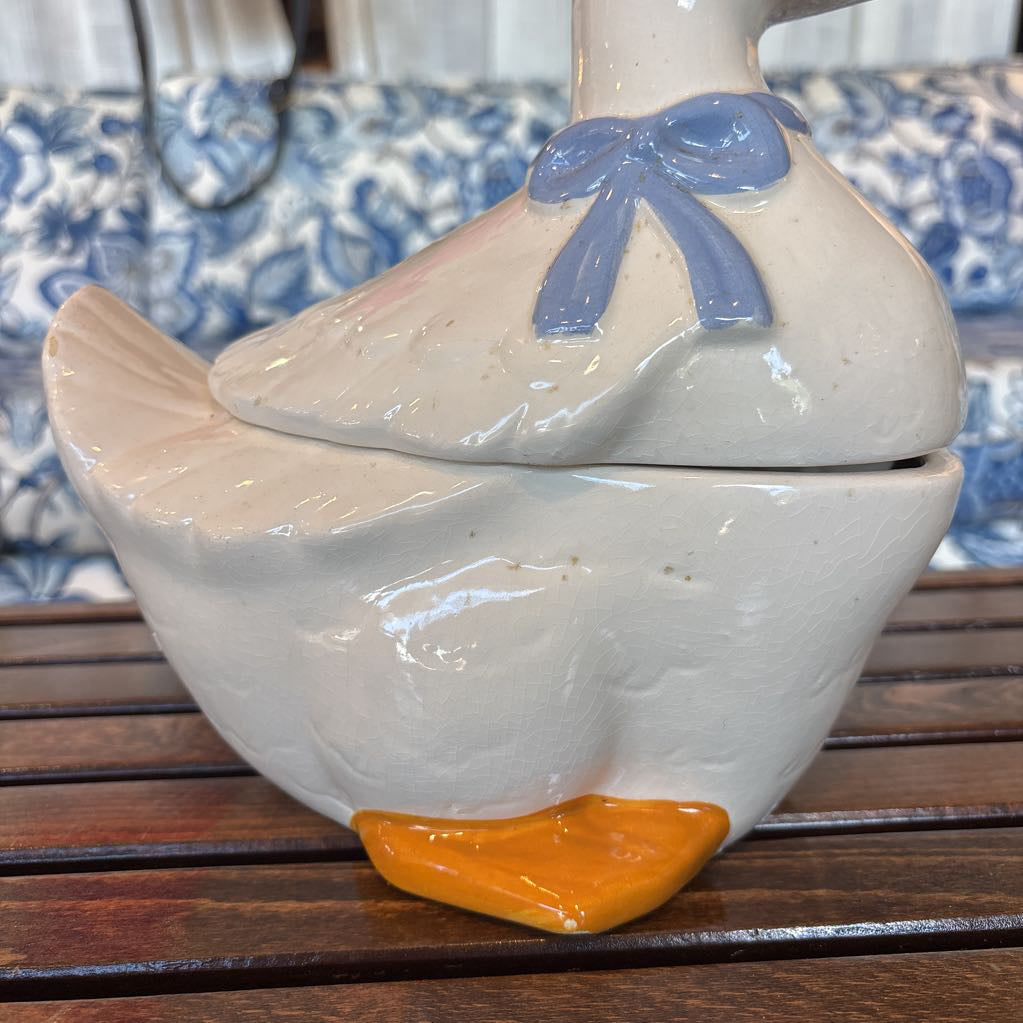 A ceramic white duck figure with a blue bow around its neck sitting on a wooden table in front of a blue and white sofa. 