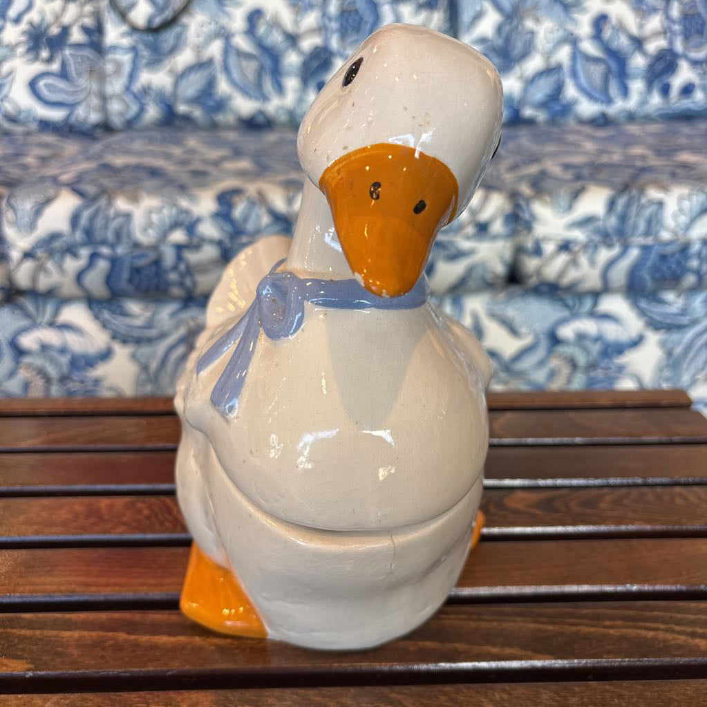 A ceramic white duck figure with a blue bow around its neck sitting on a wooden table in front of a blue and white sofa. 