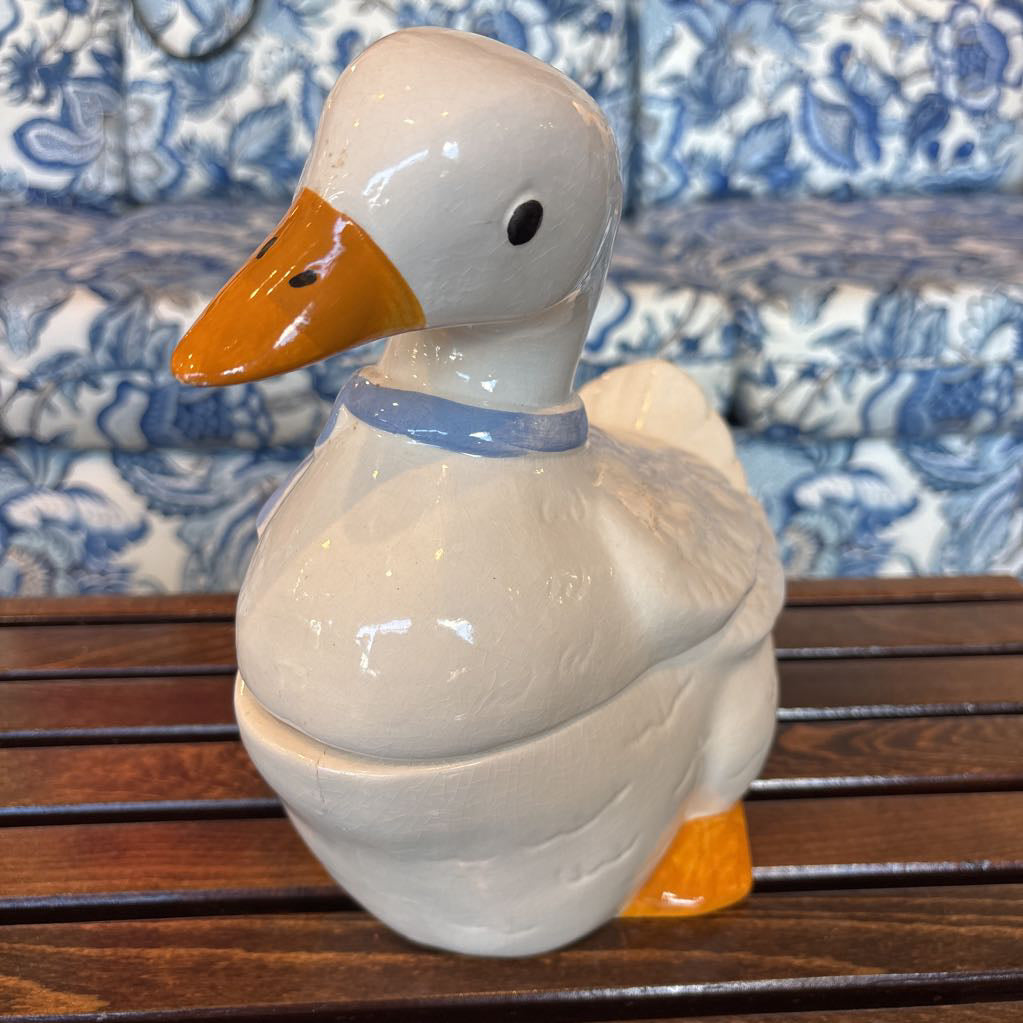 A ceramic white duck figure with a blue bow around its neck sitting on a wooden table in front of a blue and white sofa. 