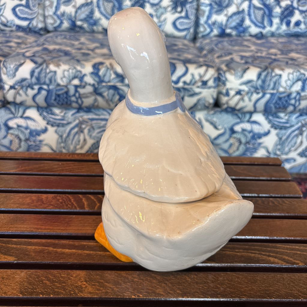 A ceramic white duck figure with a blue bow around its neck sitting on a wooden table in front of a blue and white sofa. 