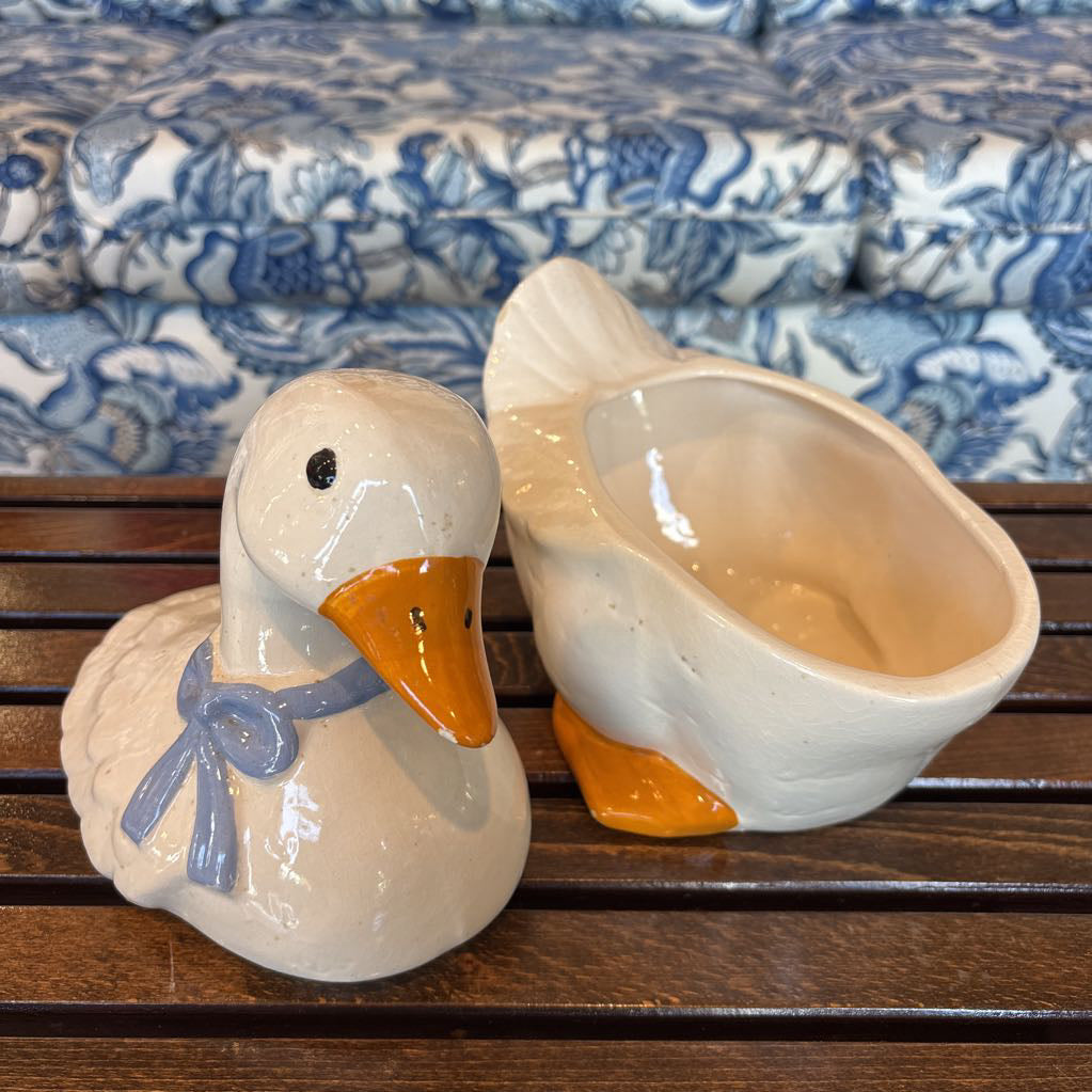 A ceramic white duck cookie jar separated into two halves sitting on a wooden table in front of a blue and white sofa. 