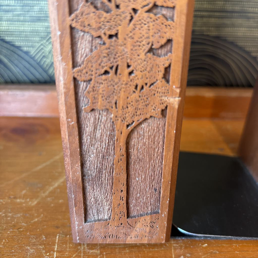 A wooden bookend with a laser cut image of a tall tree on the front sitting on a wooden surface in front of a black and gold wallpaper.