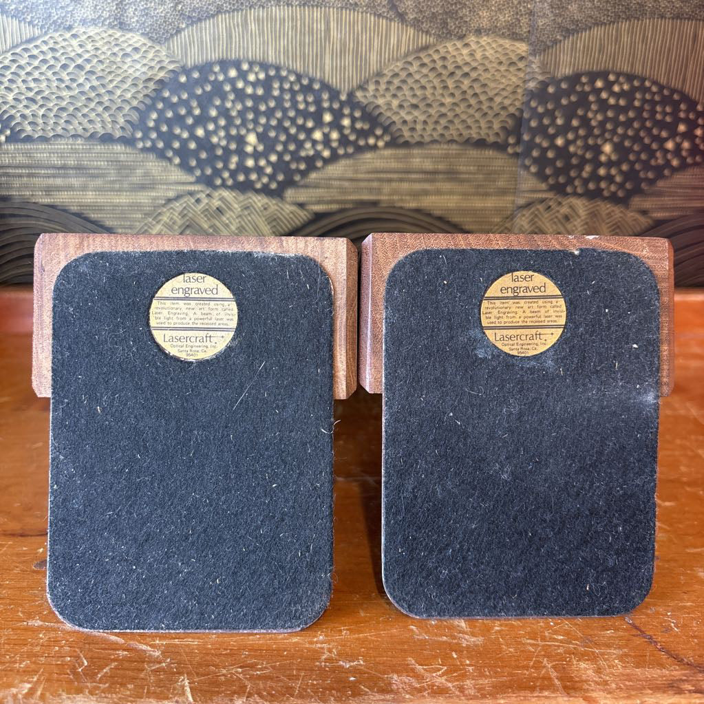 Two wooden bookends on a wooden surface in front of a black and gold wallpaper.