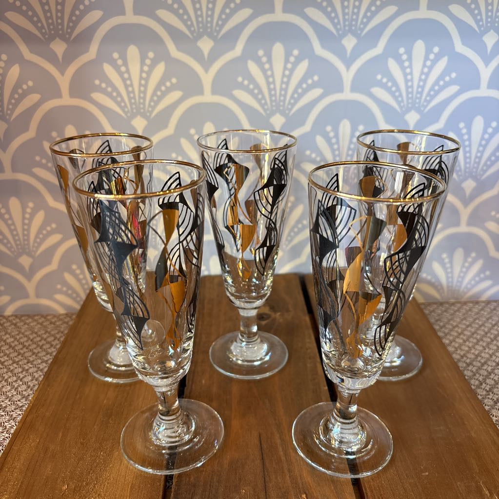 A set of 5 flared cordial glasses with black and gold wavy designs sitting on a wooden surface in front of a light blue and white wallpaper.