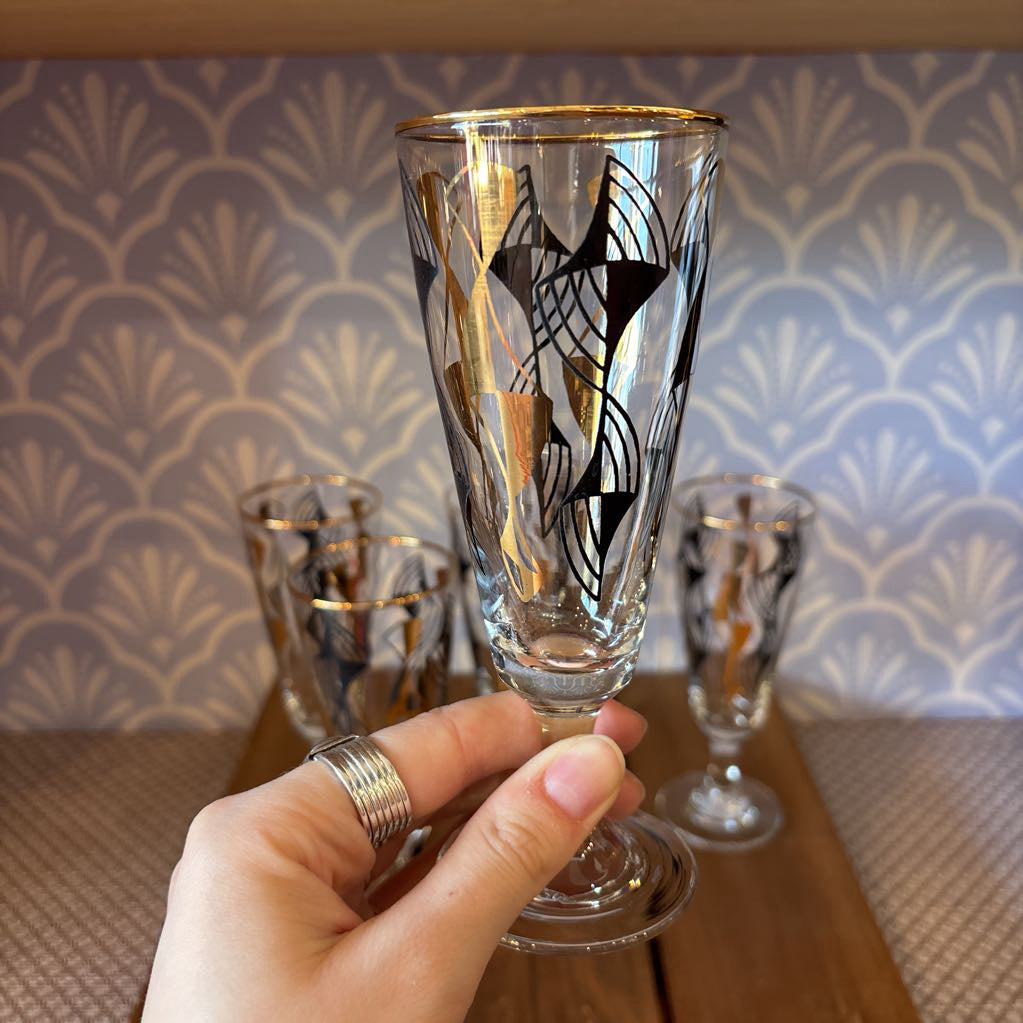 A hand holding a flared cordial glass with black and gold wavy designs in front of a light blue and white wallpaper.