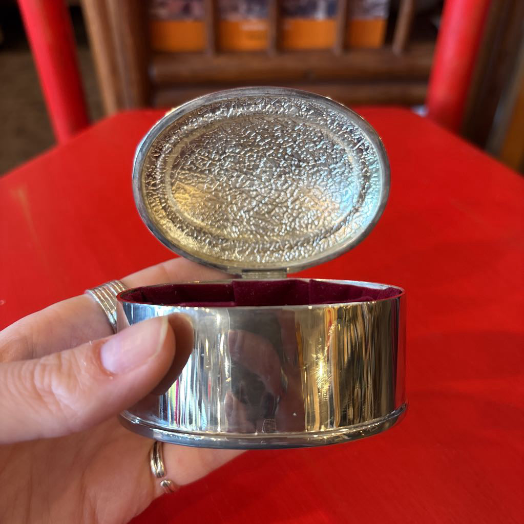 A silver jewelry box held by a hand in front of a red surface. 