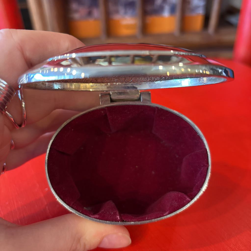 A silver jewelry box being held open by a hand showing a velvet interior and a red background. 