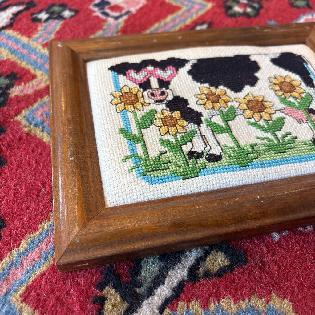 A framed cross stitch artwork of a cow with heart glasses surrounded by sunflowers. This artwork is on a red rug. 