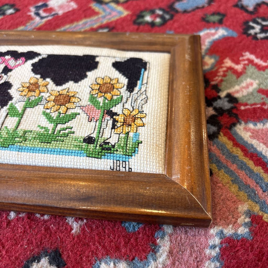 A framed cross stitch artwork of a cow with heart glasses surrounded by sunflowers. This artwork is on a red rug. 