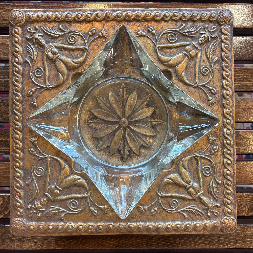 An intricate gold chalkware ashtray with designs of stylized women on the corners and a flower in the center. A glass diamond sits in the center to act as the ashtray. This whole piece sits on a wooden bench. 