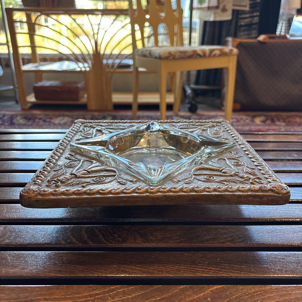 An intricate gold chalkware ashtray with designs of stylized women on the corners and a flower in the center. A glass diamond sits in the center to act as the ashtray. This whole piece sits on a wooden bench. 