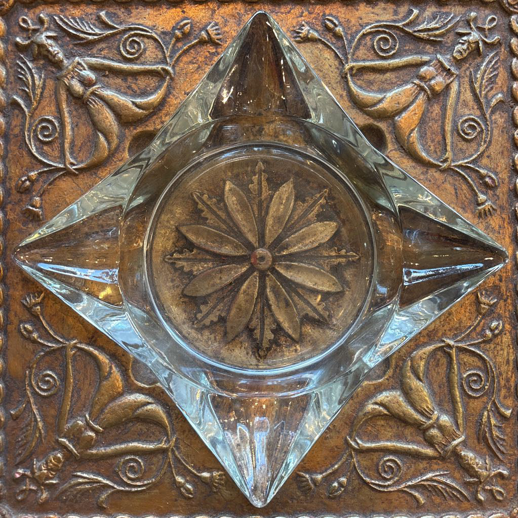 An intricate gold chalkware ashtray with designs of stylized women on the corners and a flower in the center. A glass diamond sits in the center to act as the ashtray. This whole piece sits on a wooden bench. 