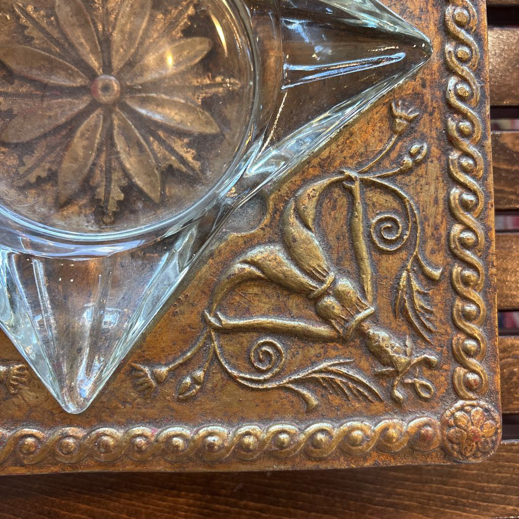 An intricate gold chalkware ashtray with designs of stylized women on the corners and a flower in the center. A glass diamond sits in the center to act as the ashtray. This whole piece sits on a wooden bench. 
