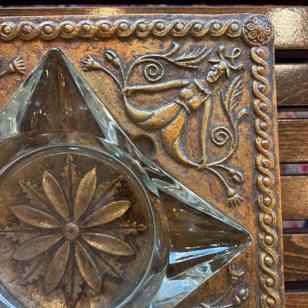 An intricate gold chalkware ashtray with designs of stylized women on the corners and a flower in the center. A glass diamond sits in the center to act as the ashtray. This whole piece sits on a wooden bench. 