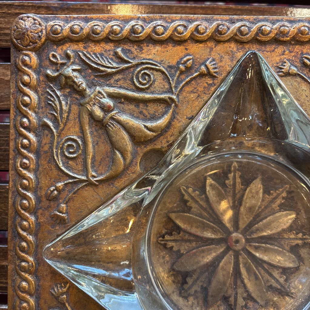An intricate gold chalkware ashtray with designs of stylized women on the corners and a flower in the center. A glass diamond sits in the center to act as the ashtray. This whole piece sits on a wooden bench. 