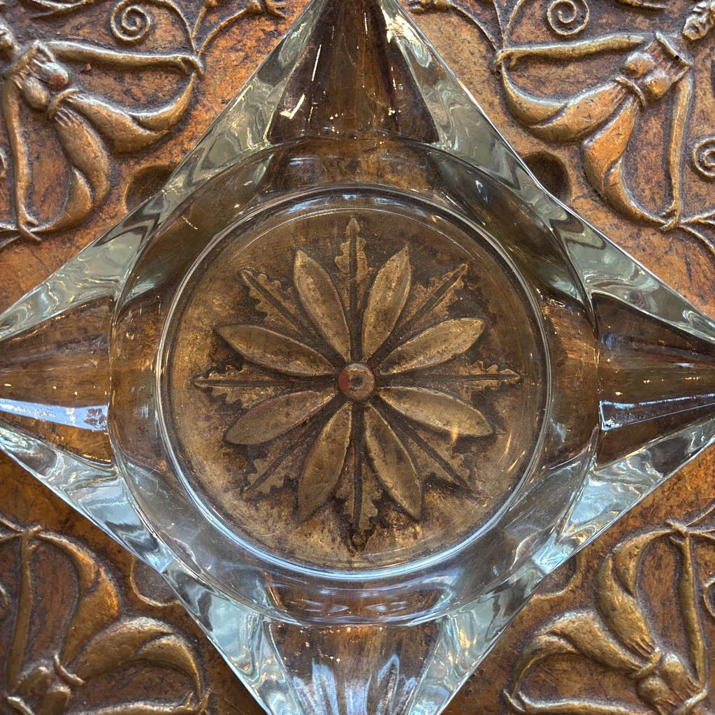 An intricate gold chalkware ashtray with designs of stylized women on the corners and a flower in the center. A glass diamond sits in the center to act as the ashtray. This whole piece sits on a wooden bench. 