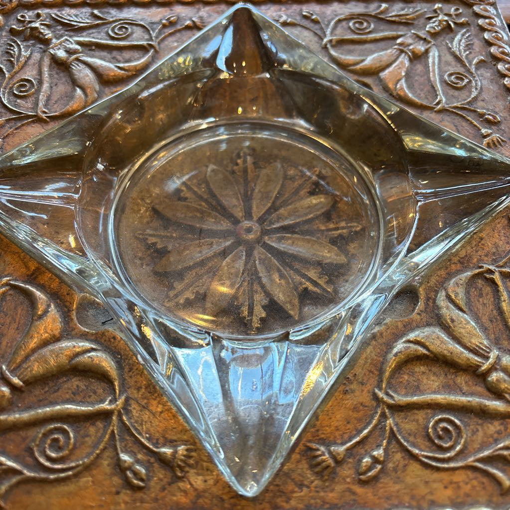 An intricate gold chalkware ashtray with designs of stylized women on the corners and a flower in the center. A glass diamond sits in the center to act as the ashtray. This whole piece sits on a wooden bench. 