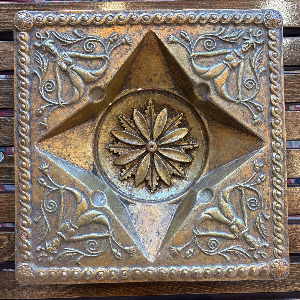 An intricate gold chalkware ashtray with designs of stylized women on the corners and a flower in the center. A glass diamond sits in the center to act as the ashtray. This whole piece sits on a wooden bench. 