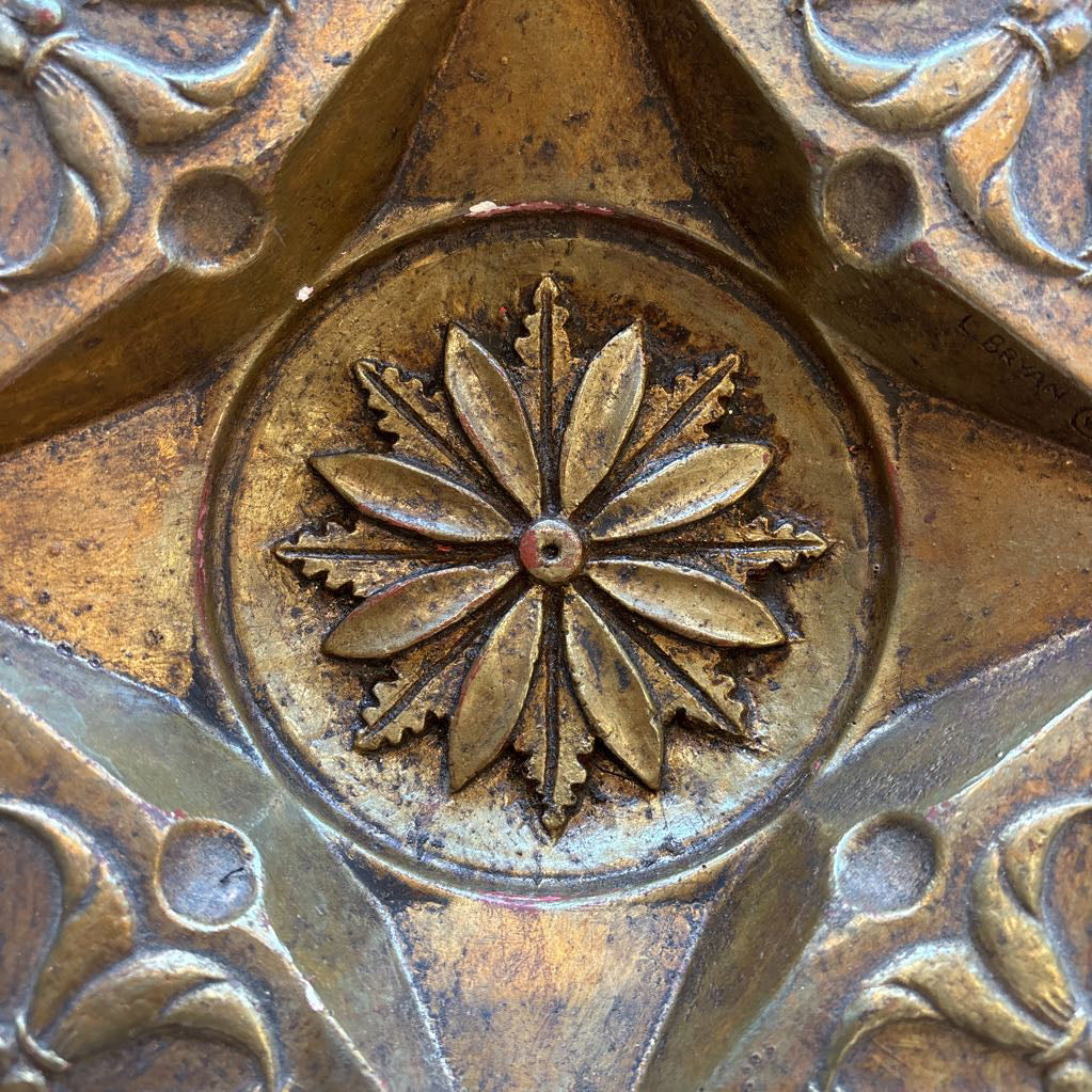 An intricate gold chalkware ashtray with designs of stylized women on the corners and a flower in the center. A glass diamond sits in the center to act as the ashtray. This whole piece sits on a wooden bench. 