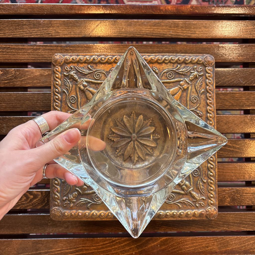 An intricate gold chalkware ashtray with designs of stylized women on the corners and a flower in the center. A glass diamond sits in the center to act as the ashtray. This whole piece sits on a wooden bench. 