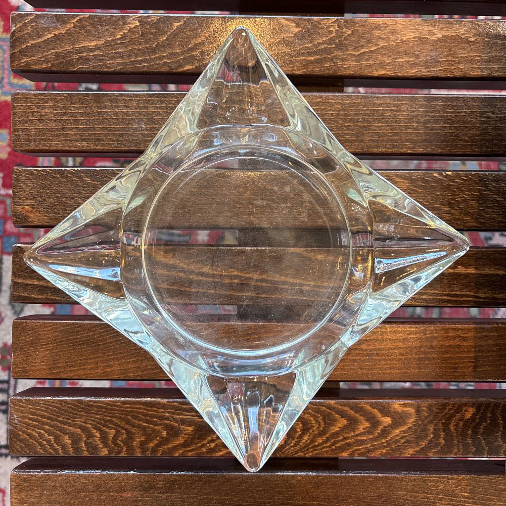 An intricate gold chalkware ashtray with designs of stylized women on the corners and a flower in the center. A glass diamond sits in the center to act as the ashtray. This whole piece sits on a wooden bench. 