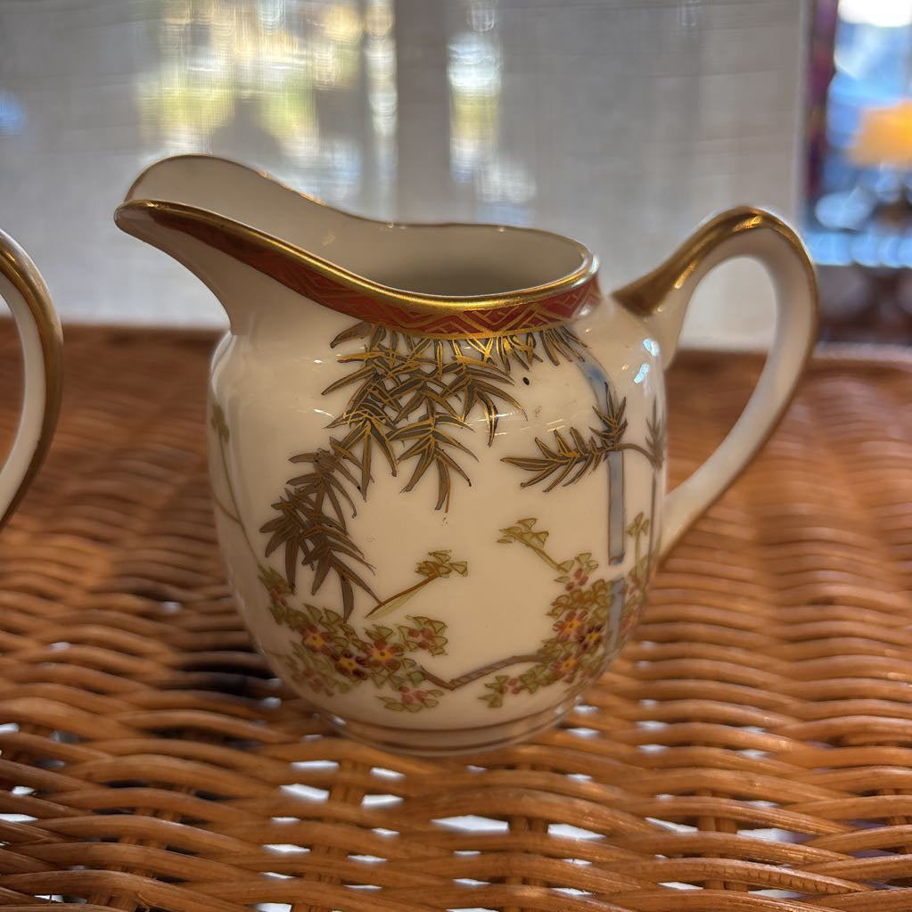 A ceramic creamer with painted floral elements on a rattan self in a room setting. 