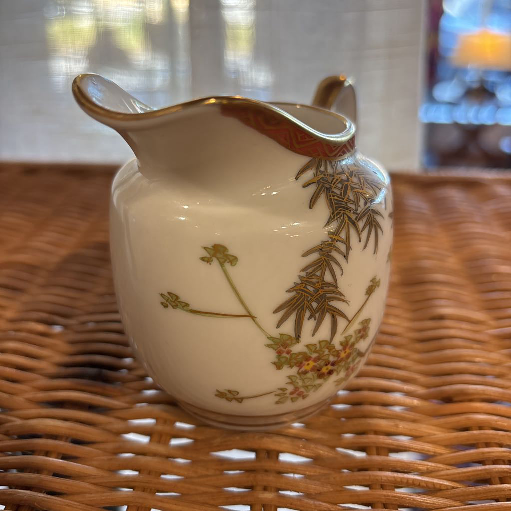A ceramic creamer with painted floral elements on a rattan self in a room setting. 