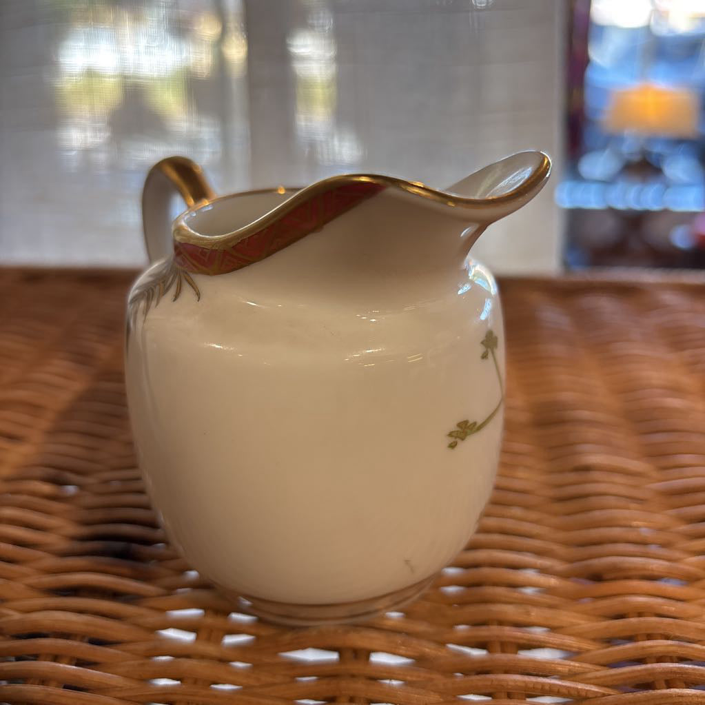 A ceramic creamer with painted floral elements on a rattan self in a room setting. 