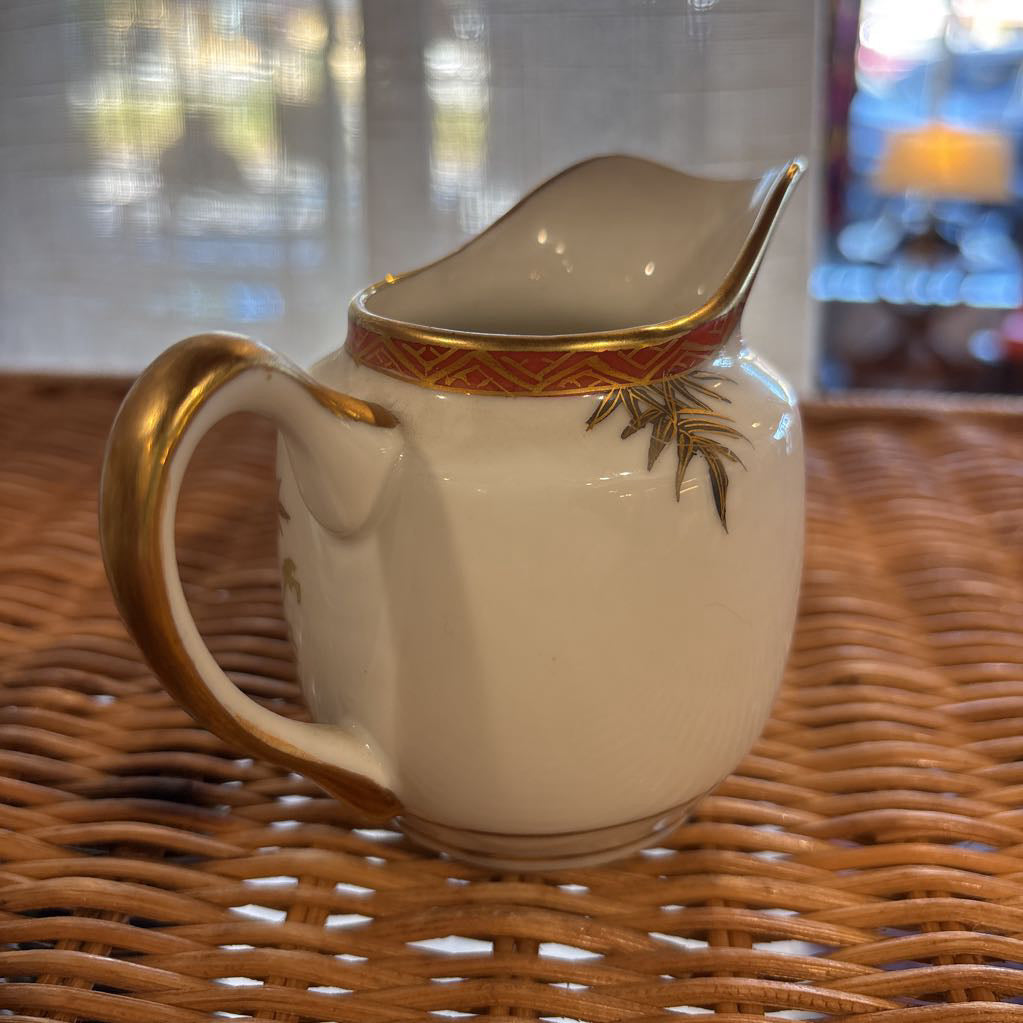 A ceramic creamer with painted floral elements on a rattan self in a room setting. 