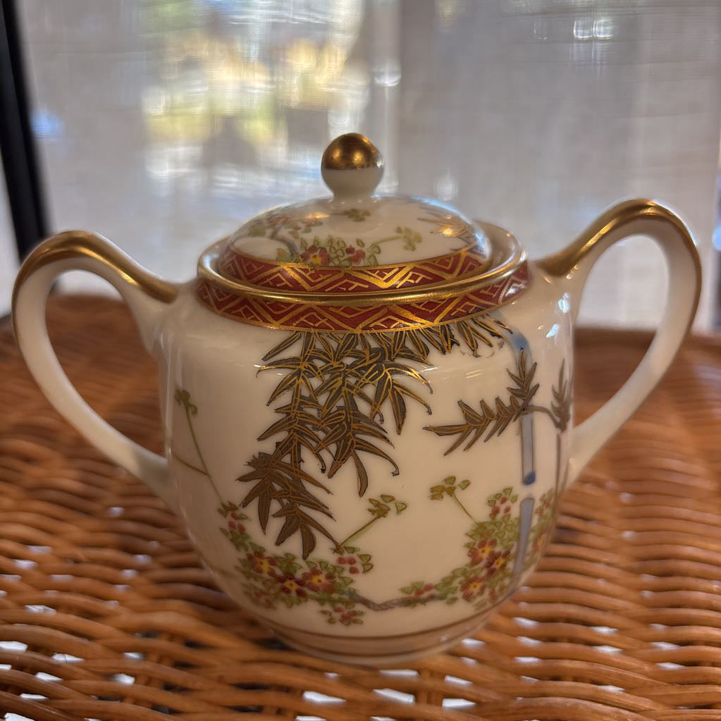 A ceramic lidded sugar bowl with painted floral elements on a rattan self in a room setting. 