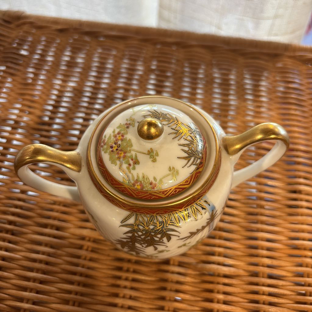 A ceramic lidded sugar bowl with painted floral elements on a rattan self in a room setting. 