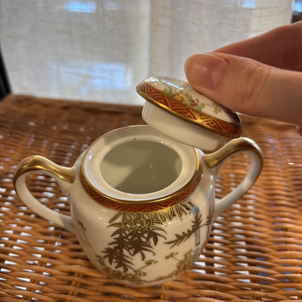 A ceramic lidded sugar bowl with painted floral elements on a rattan self in a room setting. 