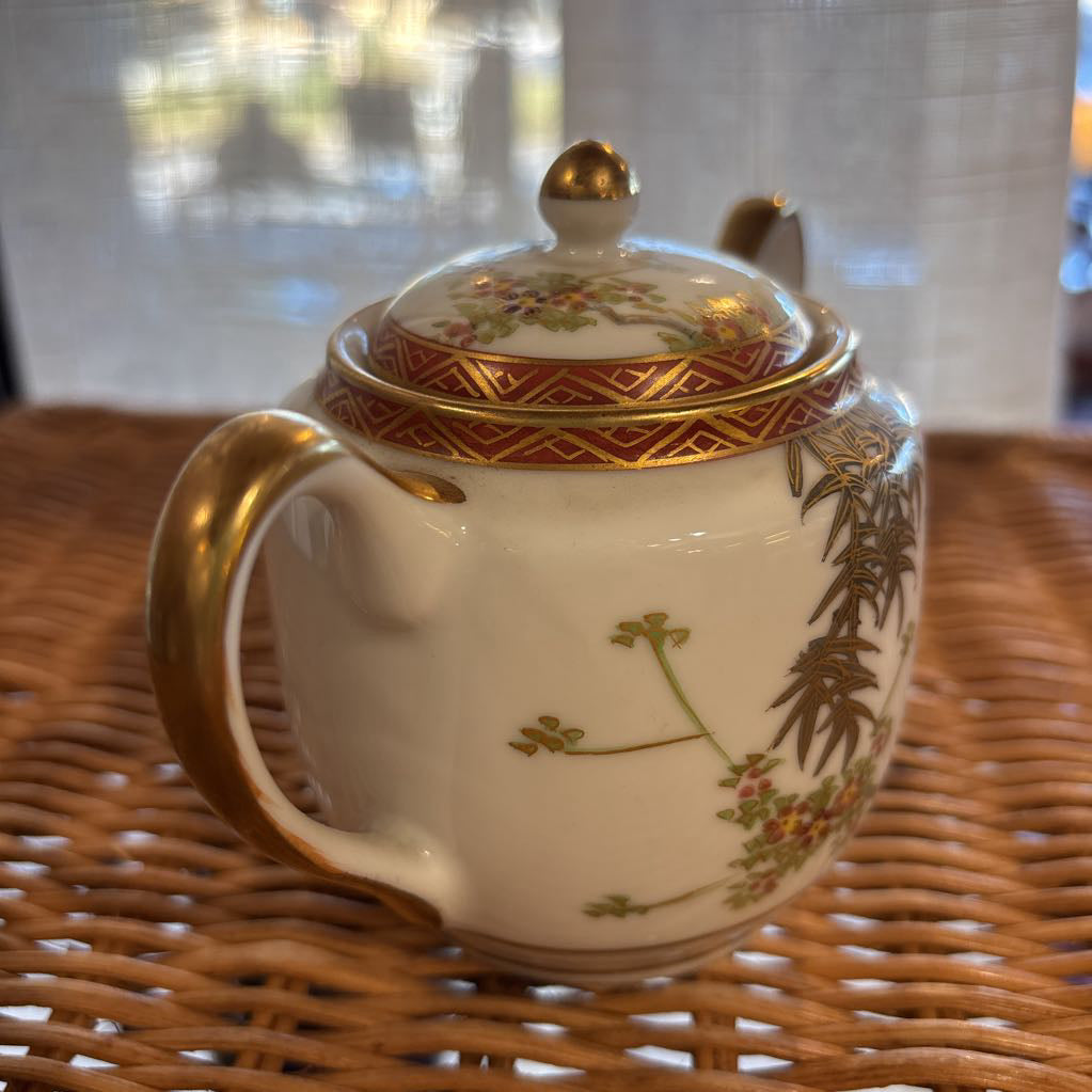 A ceramic lidded sugar bowl with painted floral elements on a rattan self in a room setting. 